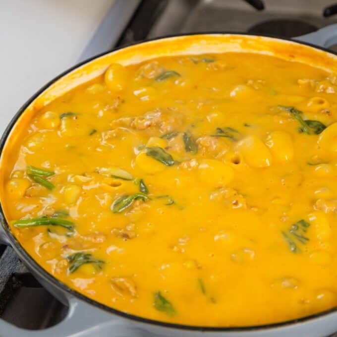 Creamy Pumpkin and Sausage Gnocchi A pot of creamy yellow macaroni and cheese with spinach and pieces of meat cooking on a stovetop.