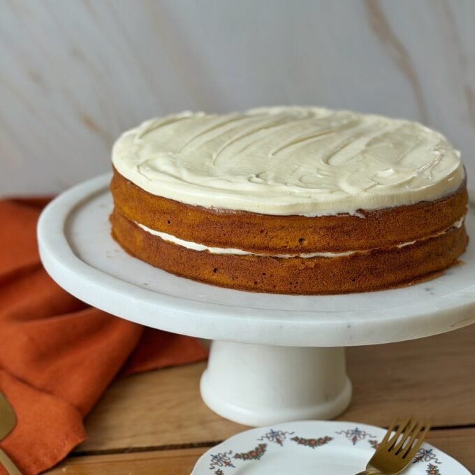 Pumpkin Spice Cake A two-layer cake with white frosting sits on a white cake stand, next to an orange cloth and a floral plate with a gold fork.