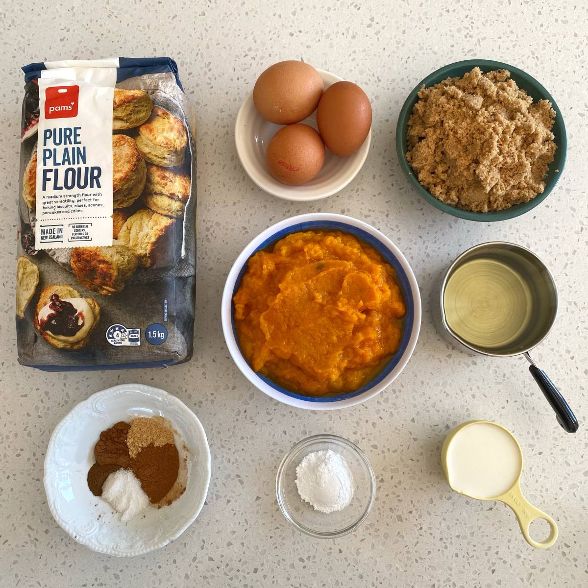 Pumpkin Spice Cake Baking ingredients on a countertop: flour, eggs, brown sugar, pumpkin puree, spices, baking powder, milk, and oil arranged in separate bowls and containers.