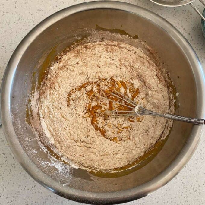 Pumpkin Spice Cake A metal bowl with flour and a yellowish liquid being mixed with a whisk on a speckled countertop.