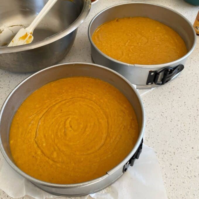 Pumpkin Spice Cake Two round springform pans filled with pumpkin cake batter sit on a countertop next to a mixing bowl and spatula.