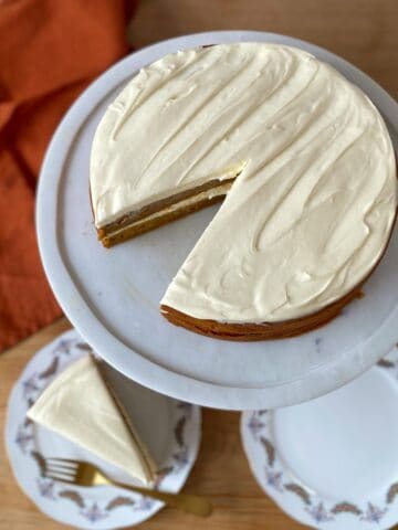 A round, frosted cake with one slice removed sits on a marble cake stand. A single slice is served on a plate with a fork nearby.