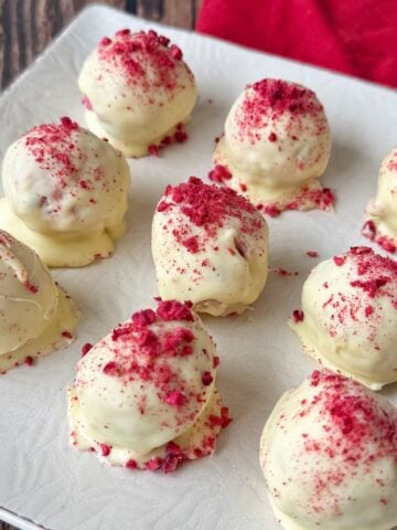 Nine white chocolate-coated truffles topped with red berry crumbs are arranged on a white square plate, with part of a red cloth visible in the background.