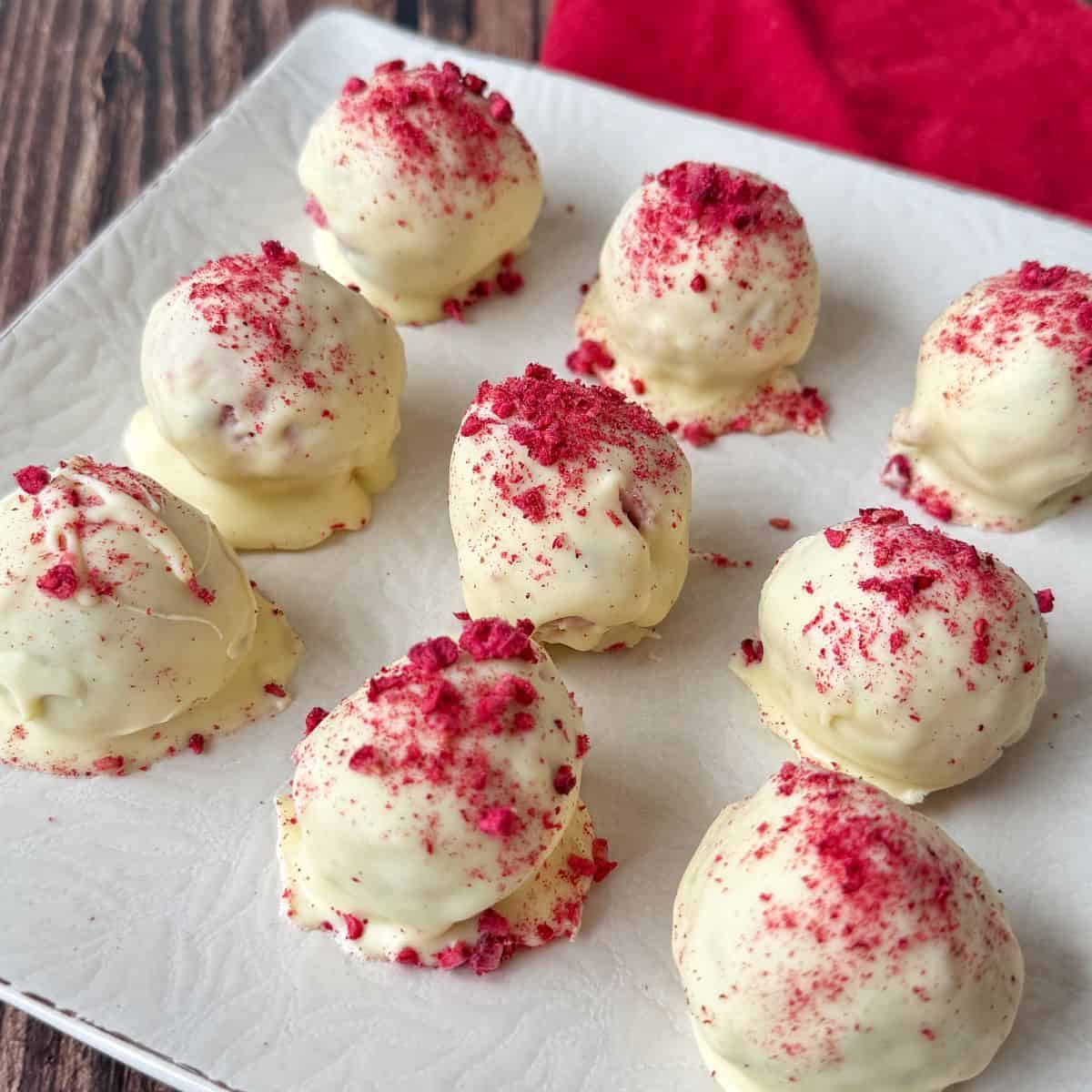 White Chocolate and Raspberry Tim Tam Balls Nine white chocolate-coated truffles topped with red berry crumbs are arranged on a white square plate, with part of a red cloth visible in the background.