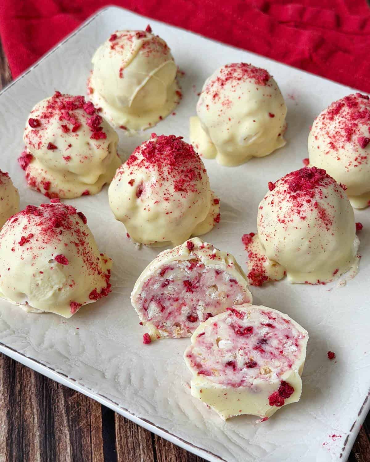 White chocolate-coated truffles topped with red crumbs are arranged on a white plate, with one truffle cut open to show a creamy filling.