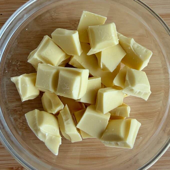 White Chocolate and Raspberry Tim Tam Balls A clear glass bowl containing several chunks of white chocolate sits on a wooden surface.