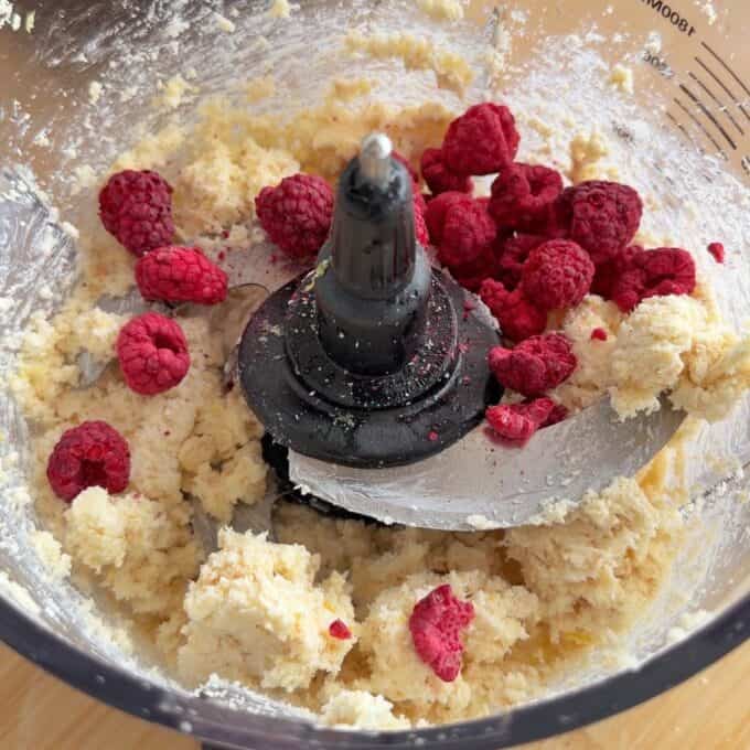 White Chocolate and Raspberry Tim Tam Balls Food processor bowl with mixed dough and whole raspberries, featuring a central metal blade and clear sides showing measurement markings.