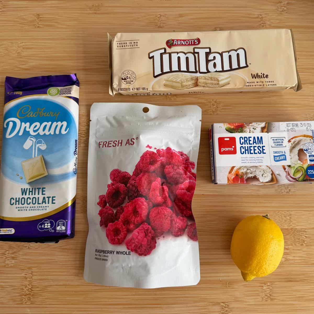 Five items on a wooden surface: a Cadbury Dream white chocolate bar, Arnotts Tim Tam White biscuits, Fresh As dried raspberries, Pams cream cheese, and a whole lemon.