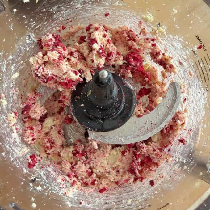 White Chocolate and Raspberry Tim Tam Balls Food processor bowl with sharp blade attachment containing a mixed blend of white and red dough ingredients, partially processed and sticking to the sides.