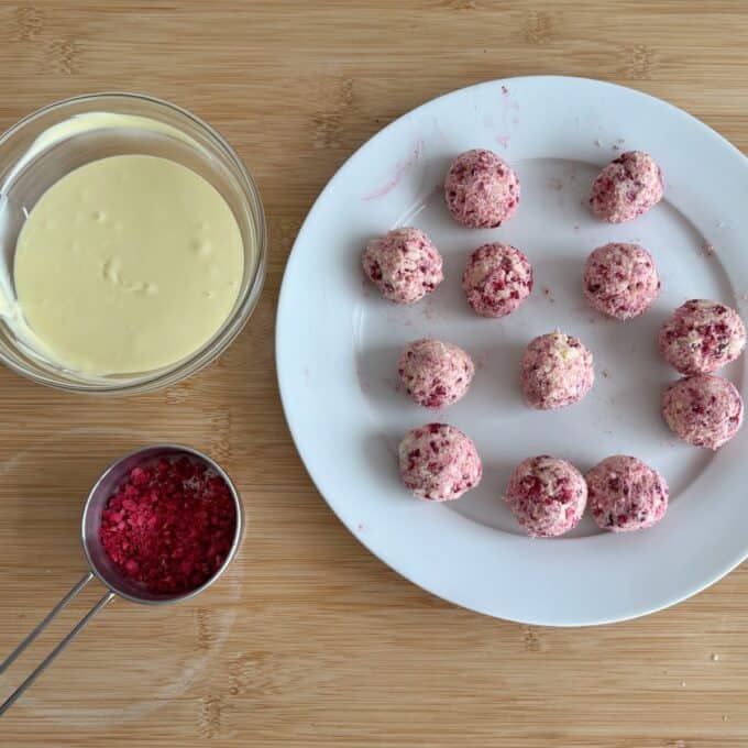 White Chocolate and Raspberry Tim Tam Balls A plate of pink speckled balls sits next to a bowl of melted white chocolate and a measuring cup of red crumbs on a wooden surface.