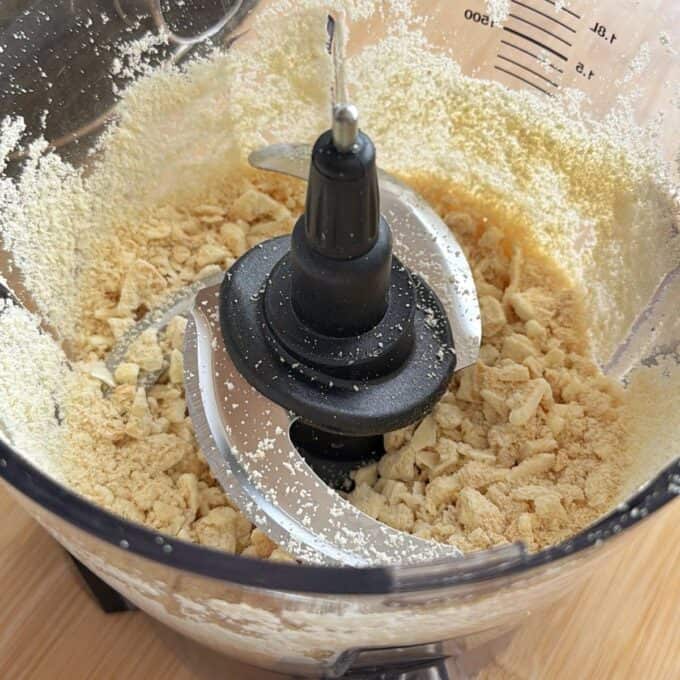 White Chocolate and Raspberry Tim Tam Balls Food processor bowl containing crushed and ground biscuit crumbs, with visible metal blades and measurement markings on the side.