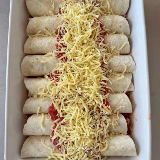 Slow Cooker Chicken Ragu A white dish contains eight rolled tortillas topped with tomato sauce and a generous layer of shredded yellow cheese, ready to be baked.