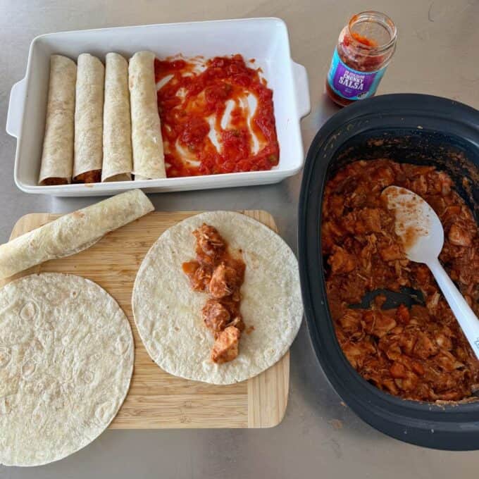 Slow Cooker Chicken Ragu Tortillas being filled with shredded chicken from a slow cooker, rolled, and placed in a dish with tomato sauce; a jar of salsa is nearby.