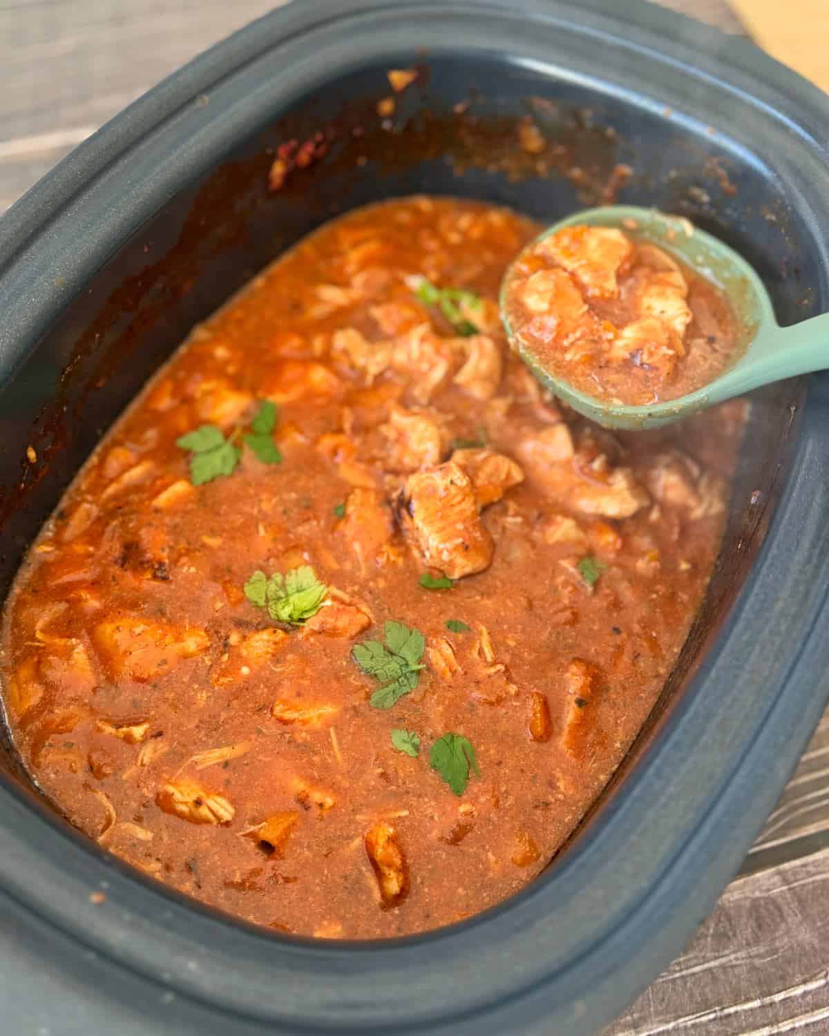 Slow Cooker Chicken Ragu A slow cooker filled with chicken curry in red sauce, garnished with cilantro, with a ladle lifting a portion.