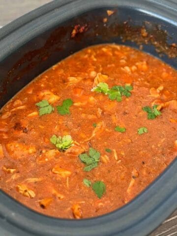 A slow cooker filled with a red tomato-based stew containing shredded chicken and garnished with fresh parsley leaves.