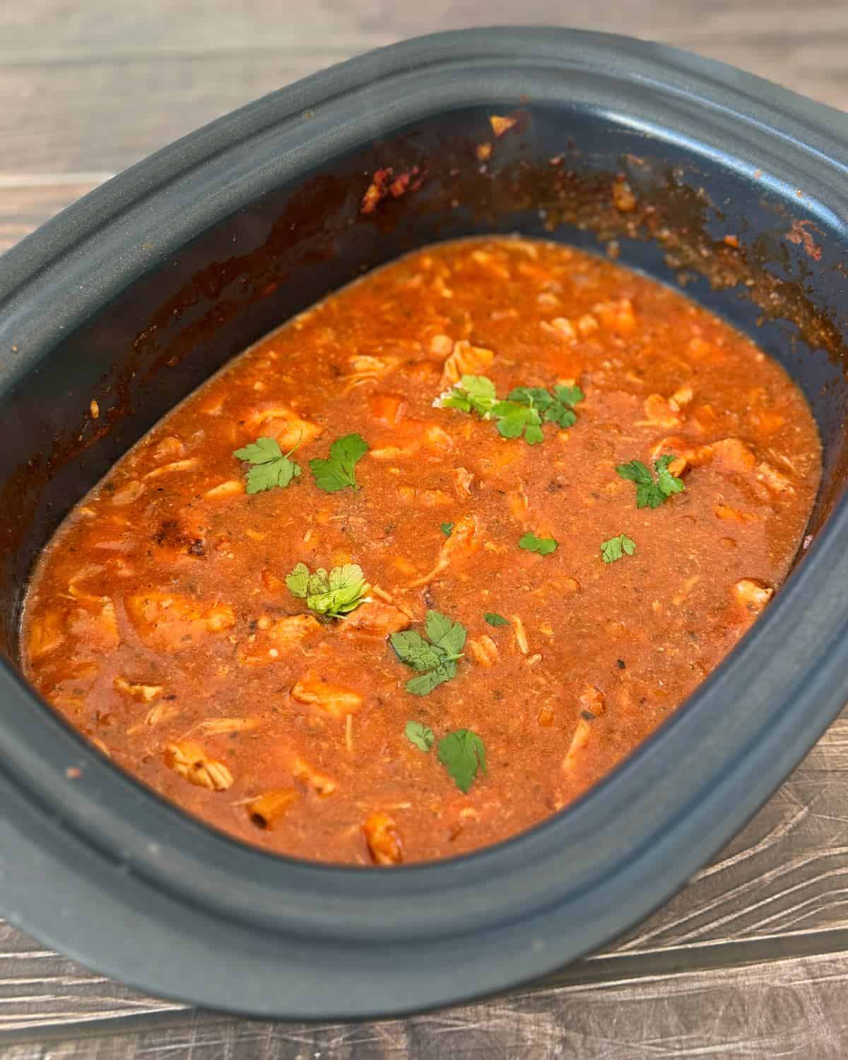 Slow cooker filled with red tomato-based stew containing shredded chicken, garnished with several sprigs of fresh cilantro.