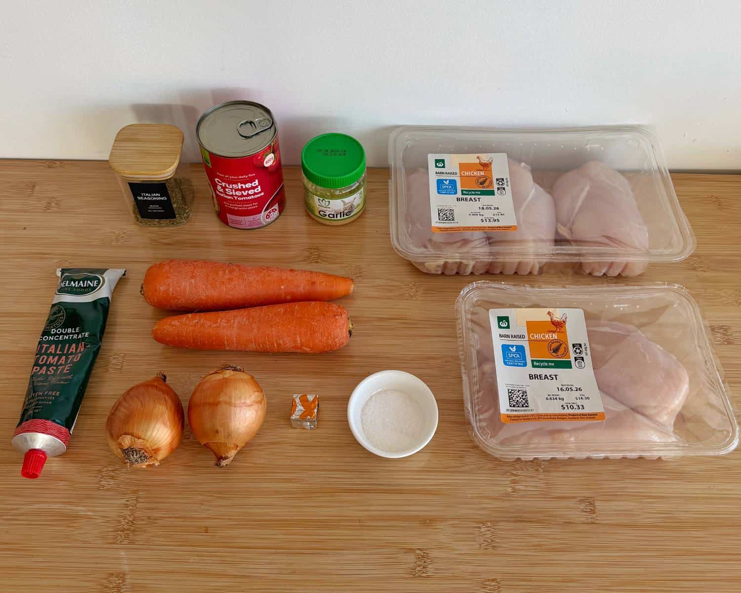 Raw chicken breasts, two carrots, two onions, tomato paste, canned tomatoes, minced garlic, salt, pepper, and a seasoning cube arranged on a wooden surface.