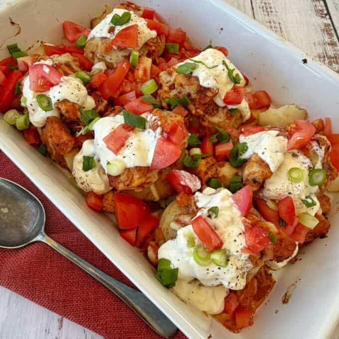 Slow Cooker Chicken Ragu Baked chicken pieces topped with chopped tomatoes, green onions, and a dollop of creamy white sauce in a white baking dish next to a spoon and red napkin.