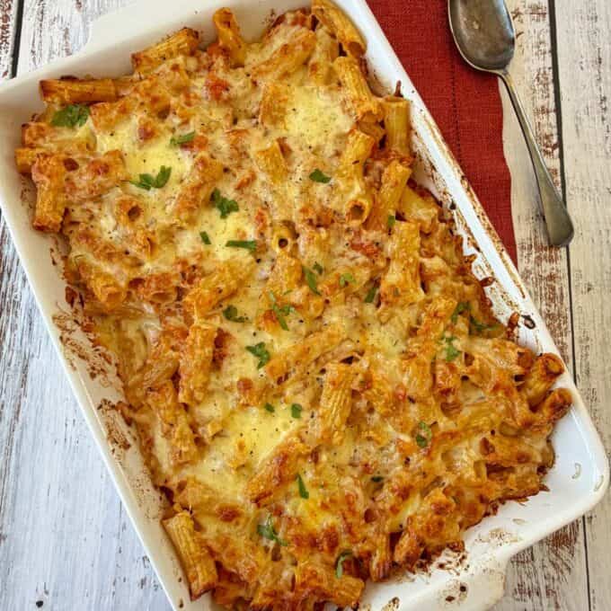 Slow Cooker Chicken Ragu Baked pasta casserole topped with melted cheese and parsley in a white rectangular dish, next to a spoon and red napkin on a wooden surface.
