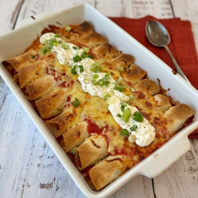 Slow Cooker Chicken Ragu A baking dish filled with baked enchiladas topped with melted cheese, sour cream, chopped green onions, and black pepper, sitting on a wooden surface with a spoon and red napkin.