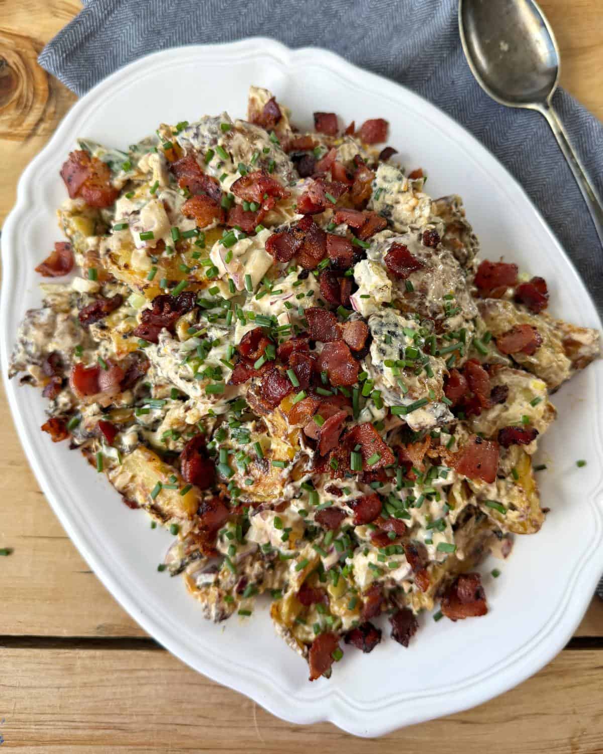 Smashed Potato Salad White platter with creamy, loaded potato salad topped with chopped bacon and chives sits on a wooden table next to a spoon and a gray napkin.