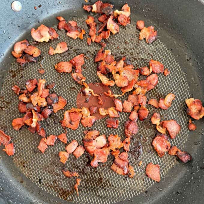 Smashed Potato Salad Crispy pieces of cooked bacon scattered in a nonstick frying pan with visible grease and browned bits.