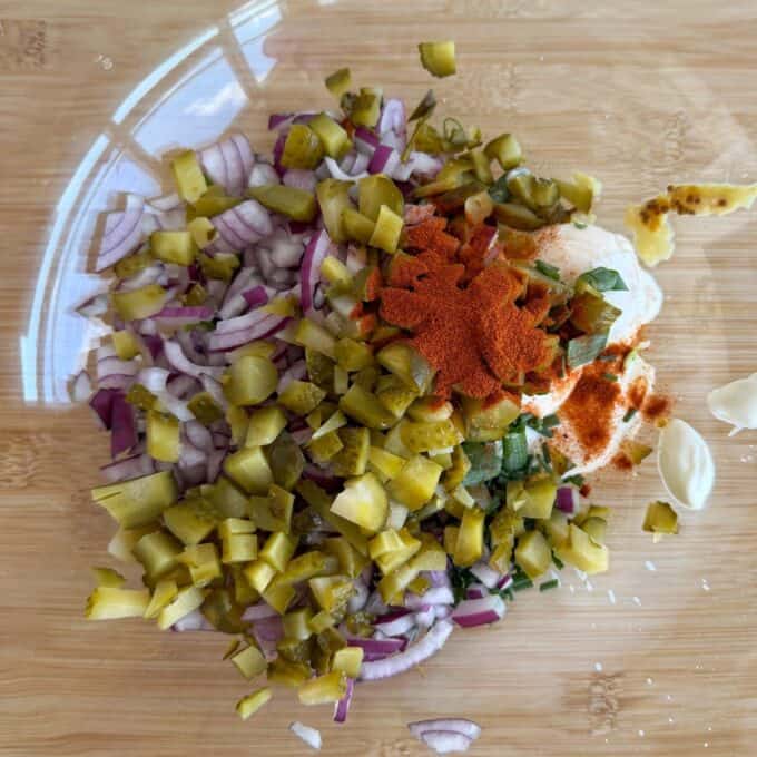 Smashed Potato Salad Chopped red onions, pickles, garlic, herbs, mayonnaise, and paprika piled on a wooden cutting board, ready to be mixed.