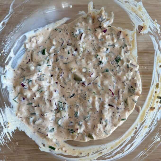 Smashed Potato Salad Bowl of creamy, mixed dip with chopped vegetables and herbs, spread unevenly on the sides of a clear glass bowl on a wooden surface.