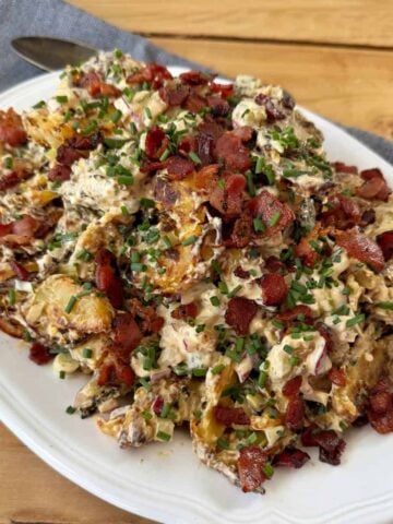 A platter of loaded potato salad topped with crispy bacon bits and chopped chives, served on a white plate with a spoon nearby.