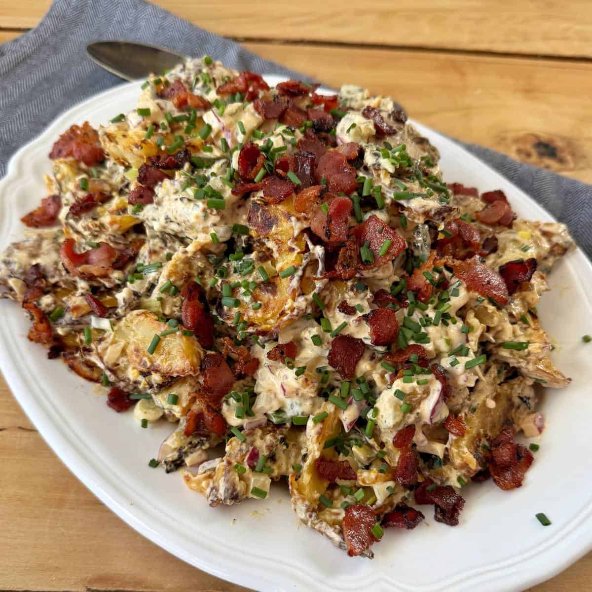 Smashed Potato Salad A platter of loaded potato salad topped with crispy bacon bits and chopped chives, served on a white plate with a spoon nearby.