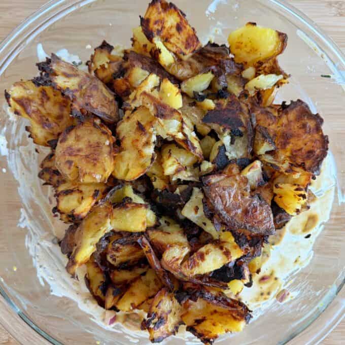Smashed Potato Salad A glass bowl filled with crispy, roasted smashed potatoes, some pieces browned and charred, on a wooden surface.