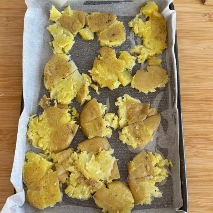 Smashed Potato Salad A baking tray lined with parchment paper holds several smashed yellow potatoes with skins, arranged in a single layer, ready for roasting.