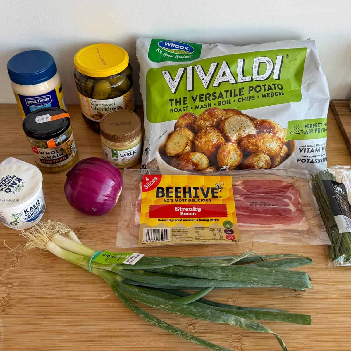 Smashed Potato Salad Various groceries on a wooden surface, including potatoes, streaky bacon, red onion, spring onions, yogurt, garlic, pickles, mustard, and cheese spread.