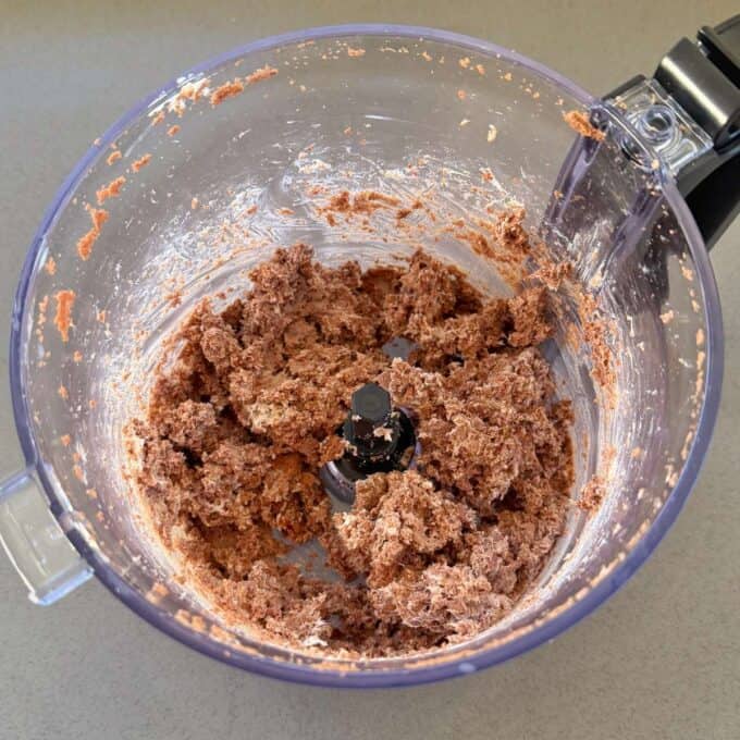 A food processor bowl containing a coarse, brownish dough mixture, partially sticking to the sides and gathered around the blade.
