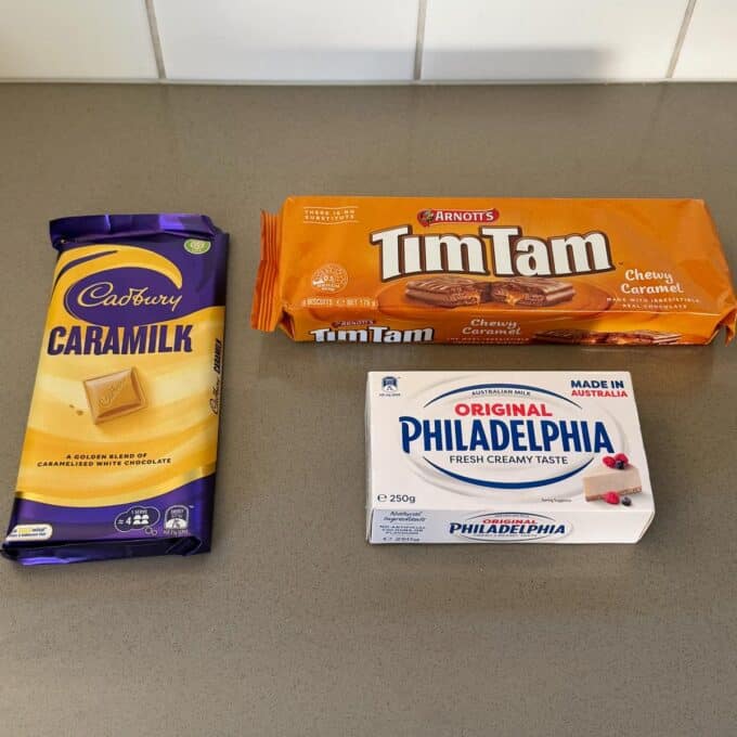 A block of Cadbury Caramilk chocolate, a packet of Arnotts Tim Tam Chewy Caramel biscuits, and a tub of Philadelphia cream cheese on a kitchen counter.