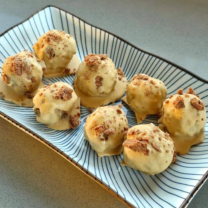 Eight round, white chocolate-coated dessert balls with crumbled toppings are arranged on a rectangular, striped plate, set on a light-colored countertop.