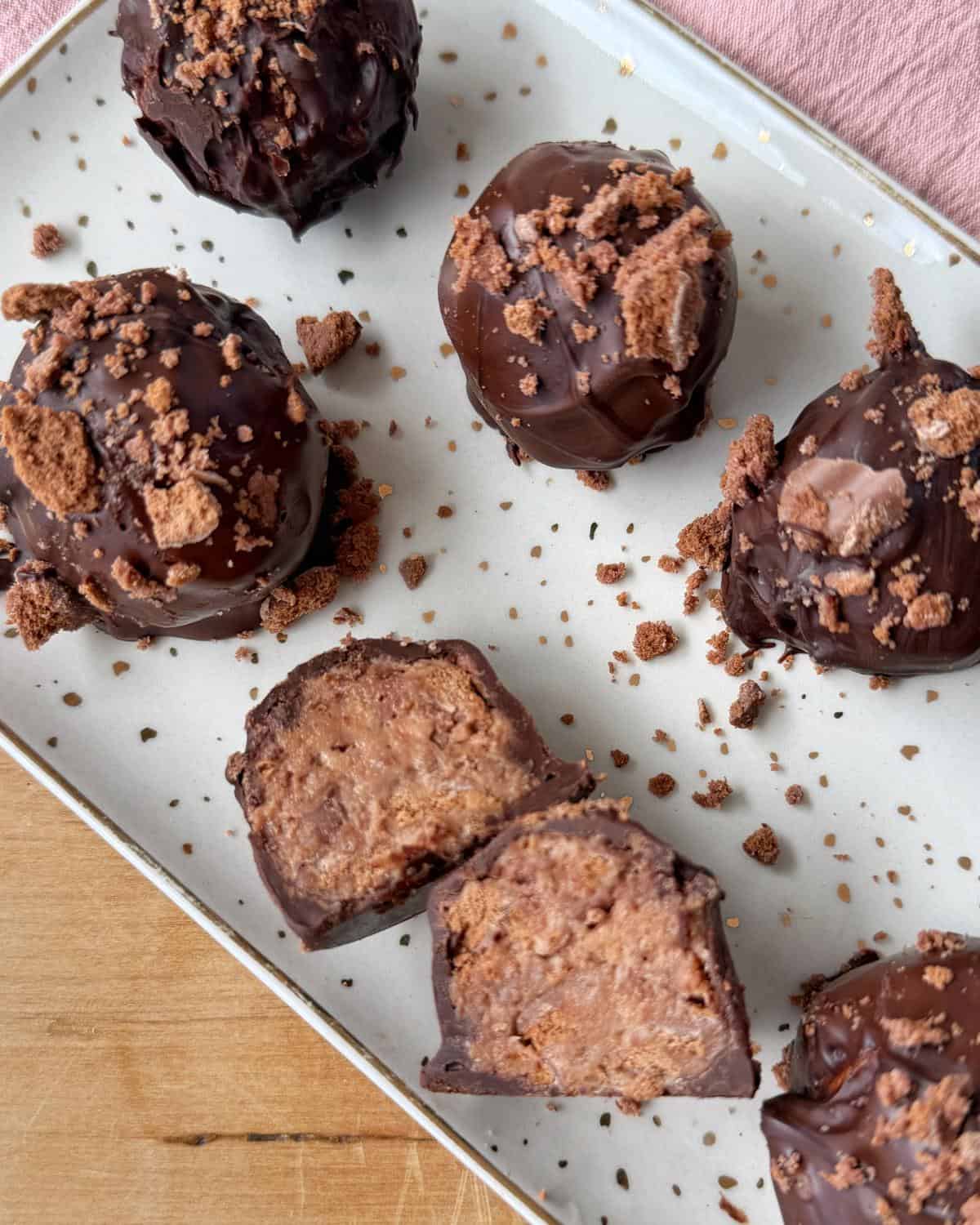 A rectangular plate with five chocolate-coated dessert balls, some topped with crumbled cookies. One ball is cut in half to show the creamy interior.