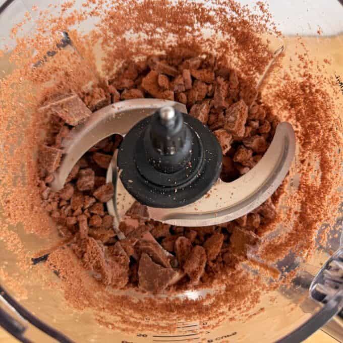 Tim Tam Cheesecake Balls Chopped chocolate pieces and cocoa powder inside a food processor with metal blades, viewed from above.