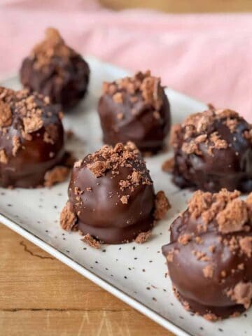 Eight chocolate truffles topped with crushed chocolate pieces are arranged on a rectangular white plate, with a pink cloth in the background.