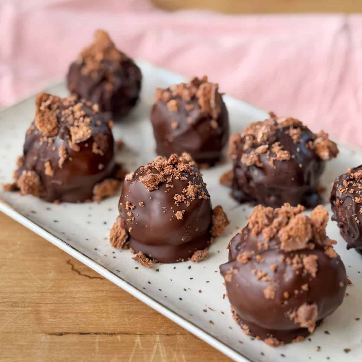 Tim Tam Cheesecake Balls Eight chocolate truffles topped with crushed chocolate pieces are arranged on a rectangular white plate, with a pink cloth in the background.