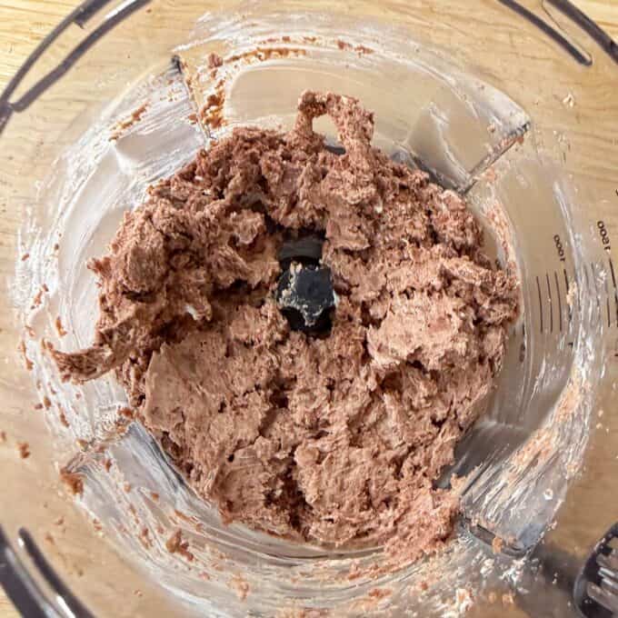 Tim Tam Cheesecake Balls A food processor bowl with a thick, light brown mixture spread around the blades, resting on a wooden surface.