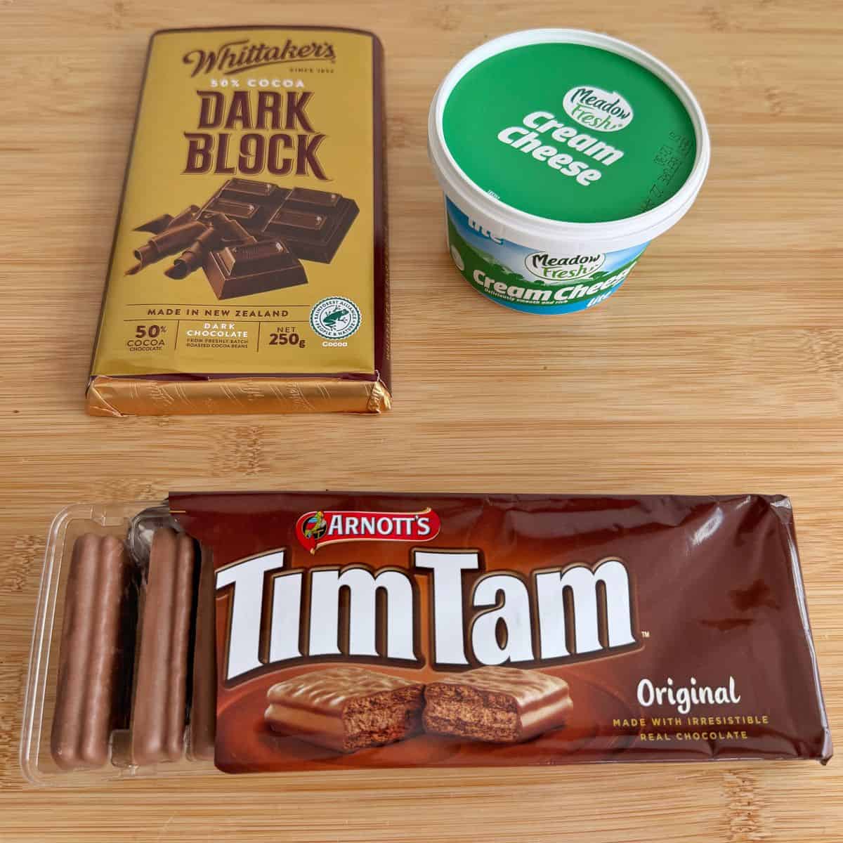 A bar of Whittakers dark chocolate, a tub of Meadow Fresh cream cheese, and a pack of Arnotts Tim Tam biscuits on a wooden surface.