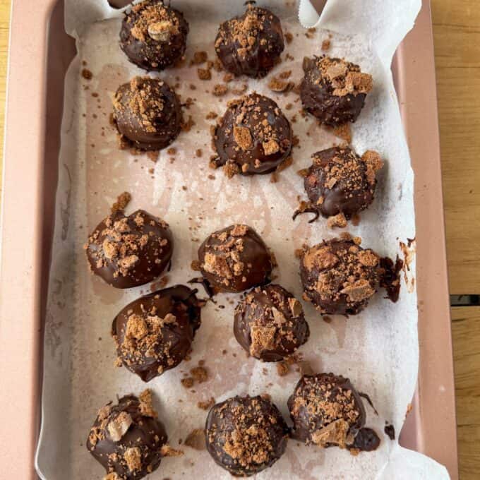 Tim Tam Cheesecake Balls Fourteen chocolate-coated truffles topped with crushed cookies are arranged on a parchment-lined baking tray.