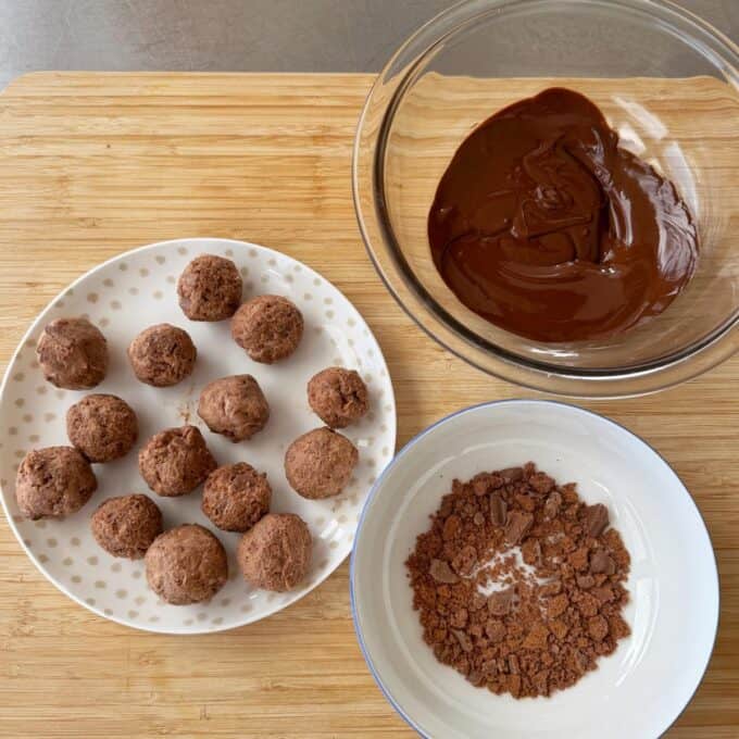 Tim Tam Cheesecake Balls A plate of chocolate truffles, a bowl of melted chocolate, and a bowl of crushed chocolate on a wooden surface.