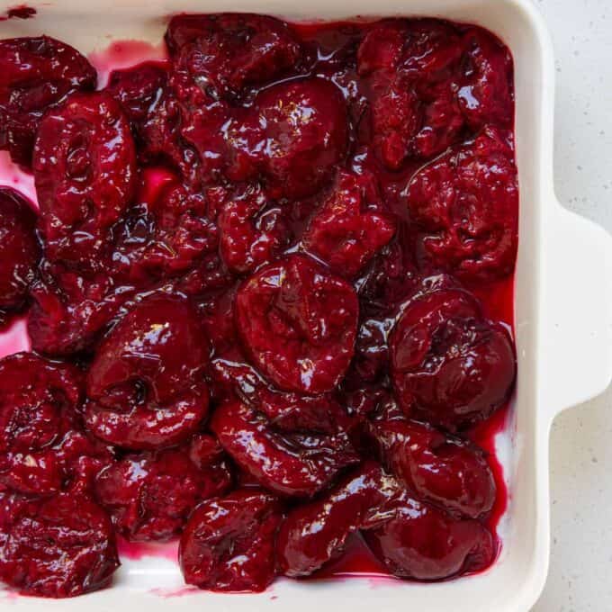 Tinned Plum Sponge Pudding A white baking dish filled with halved, cooked plums in a glossy, thick red sauce.