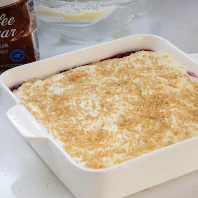 Tinned Plum Sponge Pudding A white baking dish filled with a dessert topped with sugar crystals sits on a counter near an open container and a glass bowl with remnants of mixing.