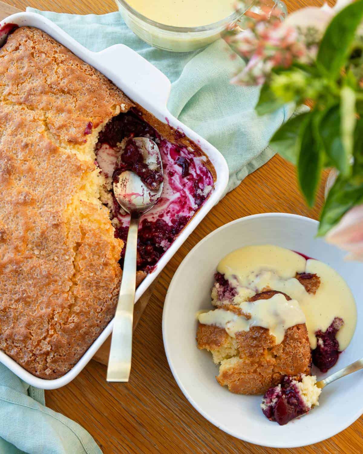 Tinned Plum Sponge Pudding A baking dish with berry cobbler and a serving spoon, next to a bowl of cobbler topped with cream sauce, set on a wooden table with a green cloth and flowers.
