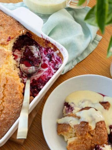 A dish of baked fruit cobbler with a serving spoon, next to a bowl of cobbler topped with custard, on a wooden table with a green napkin and vase of flowers.