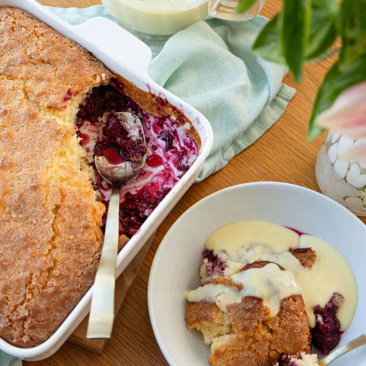 Tinned Plum Sponge Pudding A dish of baked fruit cobbler with a serving spoon, next to a bowl of cobbler topped with custard, on a wooden table with a green napkin and vase of flowers.