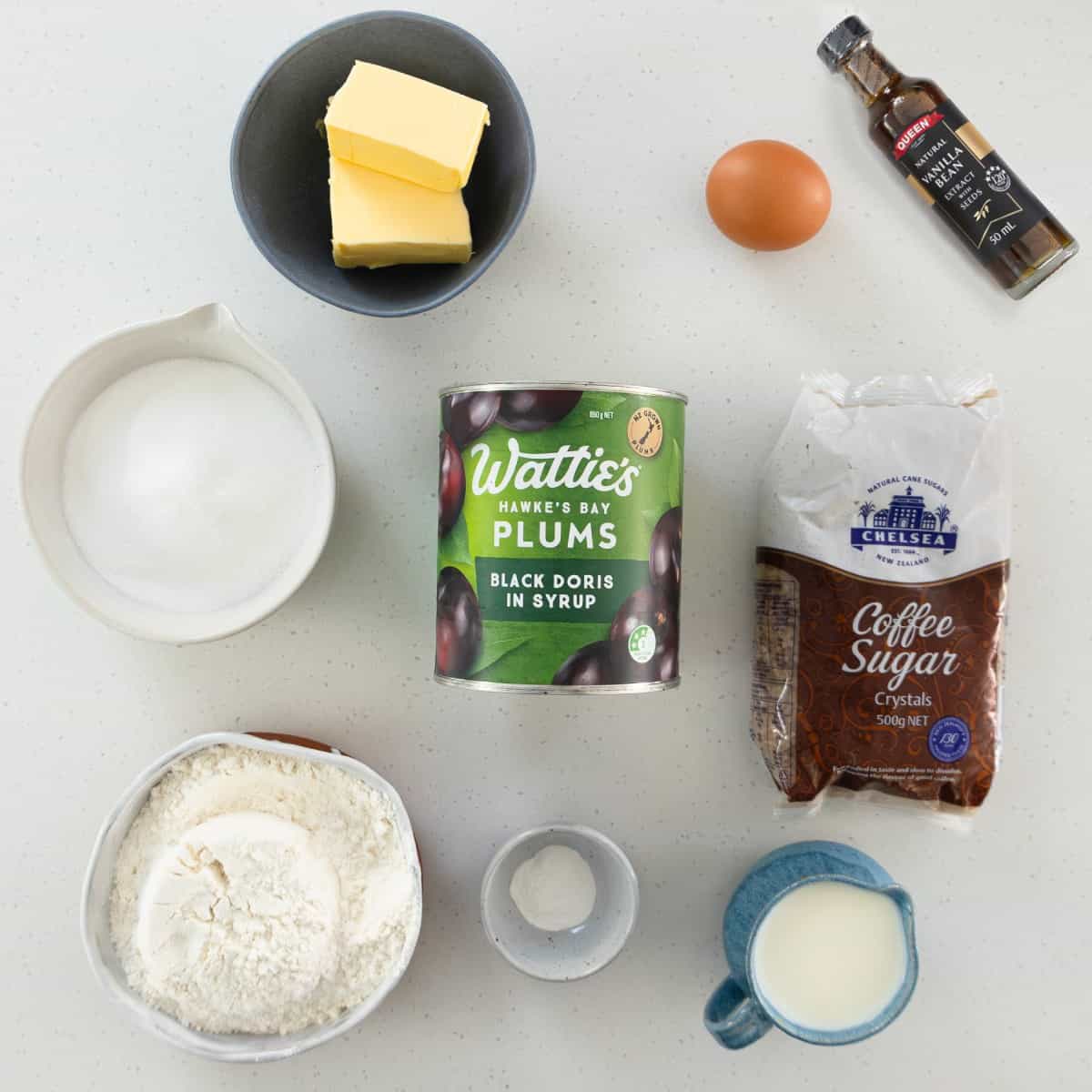 Tinned Plum Sponge Pudding Baking ingredients on a white surface: butter, egg, vanilla extract, sugar, plums in syrup, coffee sugar, flour, baking powder, and milk in separate containers.